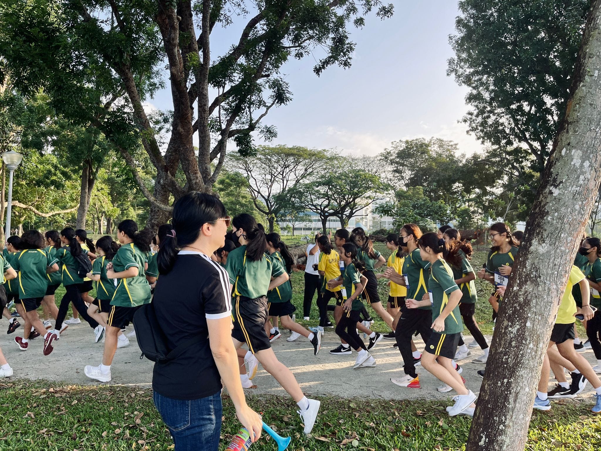 A large group of students in green and yellow uniforms running along a tree-lined path outdoors.
